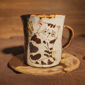 May include: Ceramic mug with a hand-painted cat illustration, complete with glasses and brown markings. The mug has a brown handle and rim, and rests on a matching saucer. The cat design includes yellow exclamation marks.