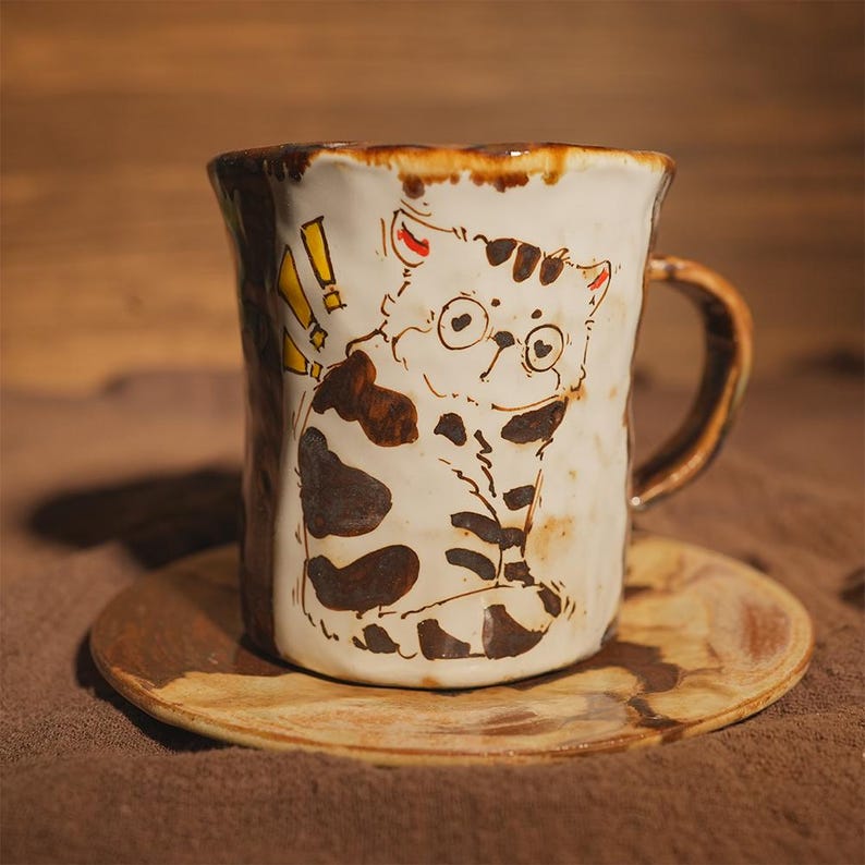 May include: Ceramic mug with a hand-painted cat illustration, complete with glasses and brown markings. The mug has a brown handle and rim, and rests on a matching saucer. The cat design includes yellow exclamation marks.