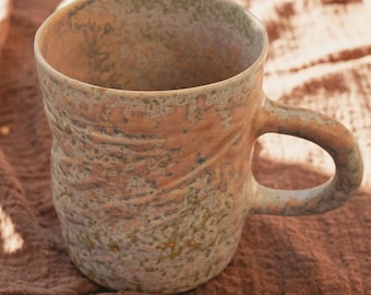 Könnte beinhalten: Handgefertigte Keramiktasse mit strukturierter Oberfläche. Die Tasse zeigt eine Mischung aus Erdtönen, darunter Pfirsich-, Braun- und Grüntöne. Sie hat einen gebogenen Griff und eine weite Öffnung, ideal für den Genuss eines Heißgetränks. Die Tasse ist ca. 10 cm hoch.