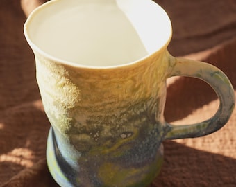 Peut inclure: Une tasse en céramique faite à la main avec une surface texturée unique. La tasse présente un dégradé de couleurs, passant d'un intérieur blanc crème à des nuances de jaune, de vert et de bleu à l'extérieur. La anse est intégrée au design.