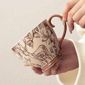 May include: A beige ceramic teacup with a brown handle and base. The cup features a detailed brown illustration of a deer, flowers, and butterflies. The cup is held by a person.