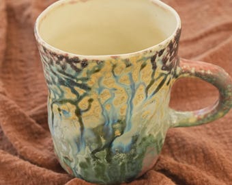 May include: A handmade ceramic mug with a unique, textured design. The mug features a blend of colors, including green, blue, yellow, and brown, with a cream-colored interior. The handle is a complementary color to the body of the mug. The mug is sitting on a brown surface.