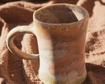 May include: A handmade ceramic mug with a curved handle. The mug has a unique, organic shape and a mottled, earthy color palette of brown, tan, and green. The mug is likely used for drinking beverages.