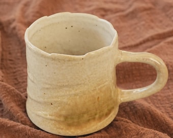 May include: A handcrafted ceramic mug with a distinctive, textured finish and a natural, earthy colour scheme. The mug has a rounded body, a robust handle, and a slightly uneven rim. The colour shifts from a creamy beige to a warm, light brown.