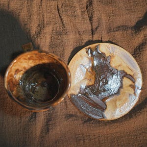 May include: A handcrafted ceramic mug and saucer set. The mug has a brown and tan marbled glaze with a small handle. The saucer features a brown and white abstract design. The set is on a brown fabric background.
