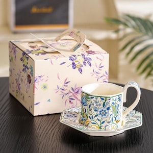Puede incluir: Un juego de taza y platillo con diseño floral, presentado con una caja de regalo a juego. La taza y el platillo tienen diseños florales azules y amarillos sobre un fondo blanco. La caja de regalo está decorada de forma similar.