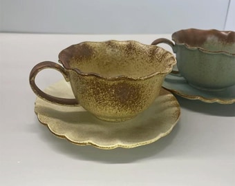 May include: Two ceramic teacups and saucers. The teacups have a scalloped rim and handle. One cup is a mottled yellow-brown, the other is green. The saucers are a matching color to the cups. The set is on a white surface.