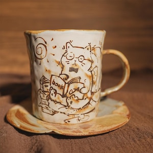 Puede incluir: Taza de cerámica color crema con una ilustración de un gato con gafas y pajarita, leyendo un libro. La taza está sobre un platillo a juego. El diseño es marrón, con detalles de sol y libro. La taza tiene un asa.