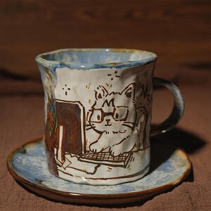 Pode incluir: Um conjunto de caneca e pires de cerâmica com um desenho de gato desenhado à mão. A caneca branca apresenta um gato de óculos trabalhando em um computador. A borda é azul e o pires é azul e marrom. A caneca tem uma alça.