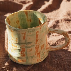 Pode incluir: Caneca de cerâmica artesanal com um design texturizado único. A caneca apresenta uma base verde clara com pinceladas laranja e verde. A alça é integrada. Uma adição perfeita para qualquer cozinha ou coleção de café.