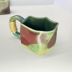 May include: A handcrafted ceramic mug with a distinctive square form and a handle wrapped in natural twine. The mug showcases a mix of green, red, and yellow hues, producing a marbled appearance. The mug is designed for hot drinks.