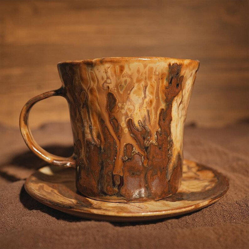 May include: A handcrafted ceramic mug and saucer set. The mug has a distinctive, textured pattern in shades of brown and cream. The handle is curved, and the saucer is round, enhancing the mug's rustic appearance.
