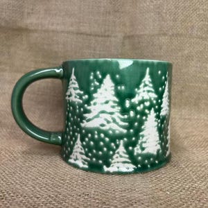 Könnte beinhalten: Eine grüne Keramiktasse mit einem weißen Weihnachtsbaum-Design. Die Tasse hat einen abgerundeten Henkel und eine glänzende Oberfläche. Die Bäume sind mit weißer Farbe detailliert, vor einem neutralen Hintergrund.