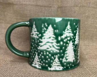 May include: A green ceramic mug with a white Christmas tree design. The mug has a rounded handle and a glossy finish. The trees are detailed with white paint and the mug is set against a neutral background.