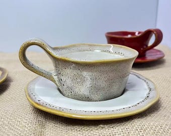 May include: A ceramic teacup and saucer set with a speckled, light brown glaze. The cup has a curved handle and sits on a matching saucer with a yellow rim. A second red teacup is visible in the background.