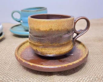 May include: A handmade ceramic coffee cup and saucer set. The cup is brown, tan, and gold with a matching saucer. A second blue cup and saucer are visible in the background. The set is on a textured surface.