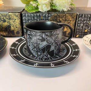 May include: Black ceramic teacup and saucer set with a white Egyptian cat design. The cup has a handle and sits on a matching saucer. The set is part of a larger collection, with boxes visible in the background.