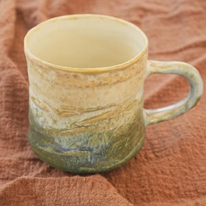 Puede incluir: Una taza de cerámica hecha a mano con un diseño único y terroso. La taza presenta un degradado de colores, pasando de un beige claro en la parte superior a un verde oscuro en la base. El asa es verde a juego. Ideal para café o té.