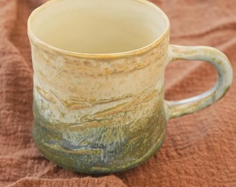 May include: A handmade ceramic mug with a unique, earthy design. The mug features a gradient of colors, transitioning from a light beige at the top to a deep green at the base. The handle is a matching green. This mug is perfect for enjoying coffee or tea.