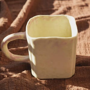 Puede incluir: Una taza de cerámica cuadrada de color amarillo pálido. La taza tiene un asa redondeada con un tono rosado. La taza está sobre una superficie marrón. Un objeto decorativo para el hogar.