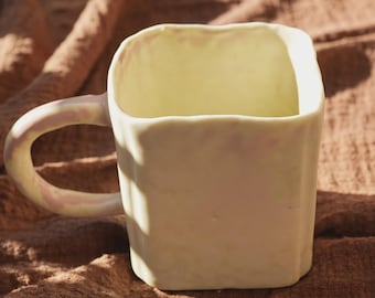 May include: A square-shaped ceramic mug in a pale yellow color. The mug has a rounded handle with a pinkish hue. The mug is sitting on a brown surface.