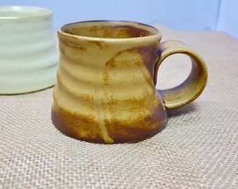 Puede incluir: Una taza de cerámica hecha a mano con un diseño texturizado y acanalado. La taza presenta una paleta de colores cálidos y terrosos con tonos marrones y beige. El asa es un lazo simple y redondeado. Otra taza es visible en el fondo.