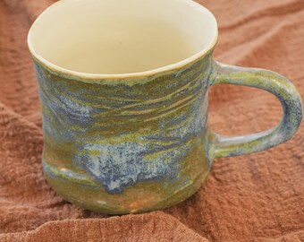 May include: A handcrafted ceramic mug with a distinctive marbled pattern. The mug has a cream-coloured interior and a textured exterior in shades of blue, green, and brown. The handle is integrated, providing a comfortable grip. Ideal for tea or coffee.