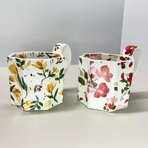 May include: Two white ceramic mugs with floral patterns. One mug features yellow tulips and green leaves, while the other displays pink flowers and red accents. Both mugs have a unique octagonal shape and curved handles.