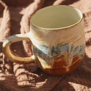 May include: A handcrafted ceramic mug with a rounded form and a substantial handle. The mug showcases a mix of hues, such as cream, brown, and blue, with a glossy surface. The inside is a pale cream shade. The mug is designed for hot drinks.