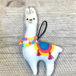 May include: A white felt llama ornament with a colorful pom-pom necklace and a saddle. The saddle is yellow, blue, and pink with red tassels. A black loop is attached for hanging. The llama is on a wooden surface.