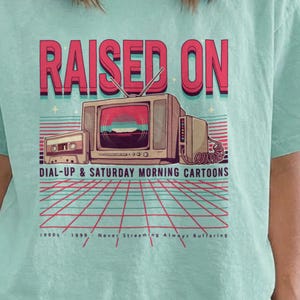 Peut inclure: Un t-shirt à manches courtes vert menthe crayeux avec une impression graphique d'une télévision vintage, d'une cassette et d'un motif en grille. Le texte indique RAISED ON DIAL-UP & SATURDAY MORNING CARTOONS, avec 1990s 1999 Never Streaming Always Buffering.