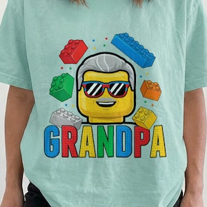 Peut inclure: Un t-shirt à manches courtes vert menthe crayeux avec un motif représentant une tête jaune de style brique avec des cheveux gris et des lunettes de soleil. Autour, des briques de construction colorées et le mot GRANDPA en lettres capitales.