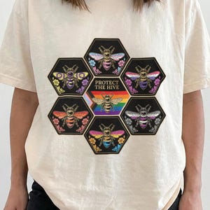 Peut inclure: Un t-shirt à manches courtes ivoire avec un motif composé de six hexagones noirs disposés en grappe. Chaque hexagone contient une abeille aux ailes colorées selon divers drapeaux de fierté. L'hexagone central montre une abeille sur un drapeau arc-en-ciel avec le texte PROTECT THE HIVE.
