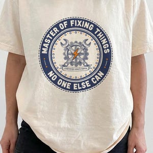 May include: An ivory short-sleeve t-shirt featuring a circular navy blue and orange graphic. The design includes a gear, two crossed wrenches, a lightning bolt, and the text "MASTER OF FIXING THINGS" and "NO ONE ELSE CAN" in a bold font.