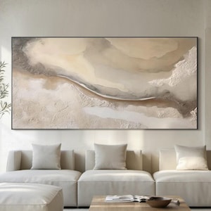 Puede incluir: Una gran pintura abstracta en tonos neutros cuelga sobre un sofá color crema. La obra de arte presenta capas texturizadas de beige, marrón y gris, enmarcadas en negro. El salón está amueblado con una mesa de centro de madera, otomanas y un jarrón alto con vegetación.
