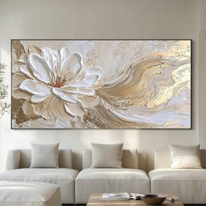 Puede incluir: Gran pintura abstracta rectangular con una flor blanca de pétalos texturizados y fondo dorado. La obra está enmarcada en negro y cuelga sobre un sofá moderno.