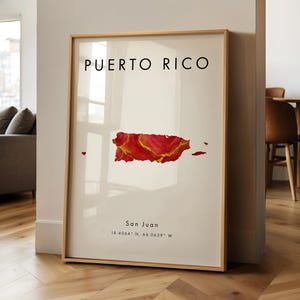 May include: A framed art print displaying the words "PUERTO RICO" above a red and gold map of the island. The city "San Juan" and coordinates are printed below the map. The print is in a gold frame and leans against a white wall.
