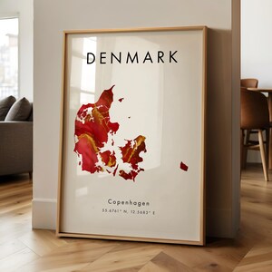 Może przedstawiać: Oprawiony wydruk przedstawiający mapę Danii z abstrakcyjnym czerwono-złotym wzorem. Słowo "DENMARK" jest wydrukowane czarnym kolorem u góry. Miasto Kopenhaga i jego współrzędne są wydrukowane u dołu. Rama jest w jasnym kolorze drewna.