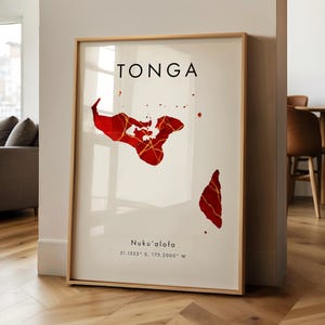 May include: A framed print showing a red map of Tonga with gold detailing. The word "TONGA" is printed in black at the top, with "Nuku'alofa" and coordinates below. The print is in a gold frame and leans against a white wall.