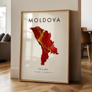 May include: Framed art print featuring a map of Moldova in red and gold hues. The word "MOLDOVA" is printed in black at the top, with "Chișinău" and coordinates below. The print is in a wooden frame.