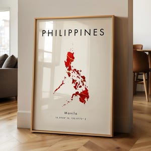 Puede incluir: Impresión artística enmarcada que muestra un mapa rojo de Filipinas con detalles dorados sobre un fondo crema. La palabra "PHILIPPINES" está impresa en negro en la parte superior, con "Manila" y coordenadas debajo. El marco es de madera clara.