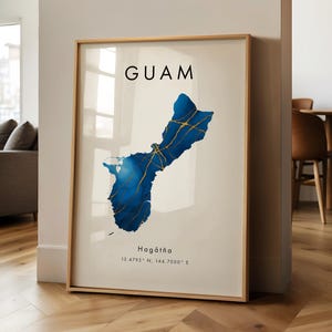 Könnte beinhalten: Gerahmter Kunstdruck mit einer Aquarellkarte von Guam in Blau- und Goldtönen. Das Wort "GUAM" ist oben in Schwarz gedruckt, darunter "Hagåtña" und Koordinaten. Der Druck befindet sich in einem hellen Holzrahmen.