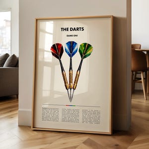May include: A framed print displaying three darts with red, blue, and green flights. The text "THE DARTS GAME ON!" is at the top. The darts have gold barrels and black shafts. The print is in a wooden frame and displayed in a room.