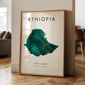 Puede incluir: Impresión artística enmarcada con la palabra "ETHIOPIA" sobre un mapa abstracto de Etiopía en verde azulado y dorado. La ciudad de Addis Abeba aparece debajo del mapa con sus coordenadas. La impresión está en un marco dorado.