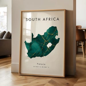 May include: A framed print of a map of South Africa, with a teal and gold design. The words "SOUTH AFRICA" are at the top, with "Pretoria" and coordinates below. The print is in a gold frame, leaning against a white wall.