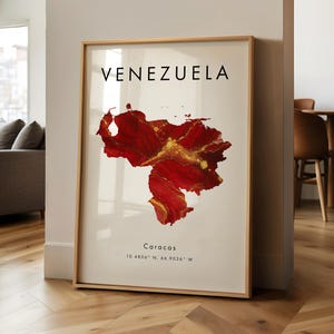 May include: A framed print of Venezuela's map, rendered in red and gold hues. The word "VENEZUELA" is printed above the map, with "Caracas" and coordinates below. The artwork is displayed in a room with wooden flooring.