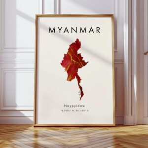 May include: A framed art print showing a map of Myanmar in red and gold hues. The country's name is printed in black at the top, with the city of Naypyidaw and its coordinates below. The print is encased in a wooden frame.