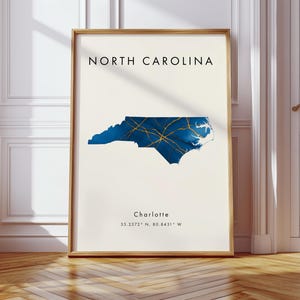 Puede incluir: Una lámina enmarcada con un mapa de Carolina del Norte en azul oscuro con líneas de carreteras doradas. El texto dice NORTH CAROLINA en la parte superior y Charlotte 35.2272 N, 80.8431 W en la parte inferior sobre un fondo crema en un marco dorado.