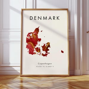 May include: A framed print featuring a map of Denmark in shades of red and gold. The word "DENMARK" is printed in black at the top, with "Copenhagen" and coordinates below the map. The frame is a light brown color.