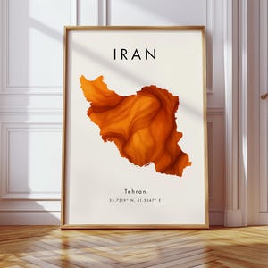 Puede incluir: Impresión artística enmarcada que presenta un mapa naranja detallado de Irán. La palabra "IRAN" está impresa en negro en la parte superior, con "Teherán" y coordenadas debajo. La obra de arte se exhibe en una habitación con paredes blancas y suelo de madera.